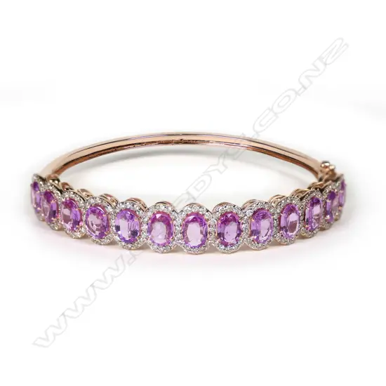 A 14ct rose gold pink sapphire and diamond halo bangle, thirteen oval cut pink sapphires (eye clean to slightly included), claw set, each surrounded by a halo of round brilliant cut diamonds (J-M/champagne colour, SI1-I2 clarity), hinged closure with safety catch, TSW 10.27ct, TDW 1.98ct.