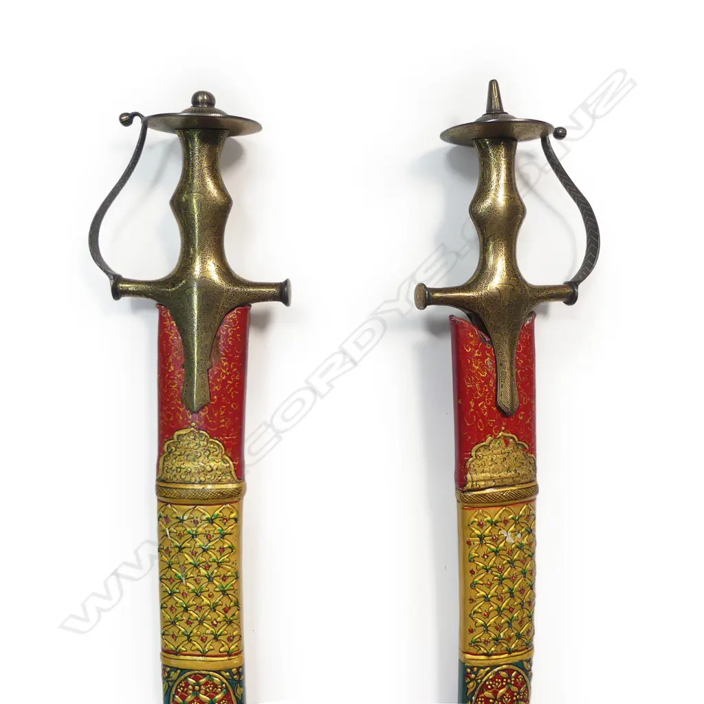 Two Tulwar Swords and matching Dahl Shield from the Punjab Region, India. A matching pair of 28" deeply-curved wootz blades with a gold cartouche to each; double-edged tips and serrated notch-backed to 3/4 of their lengths. The hilts are profusely gold damascened (one with a pommel spike, the other a dome). Matched leather scabbards with raised gilding on a red ground. Along with a matching Dahl shield, dia.20". Believed to have been made as a presentation set. Image 1++