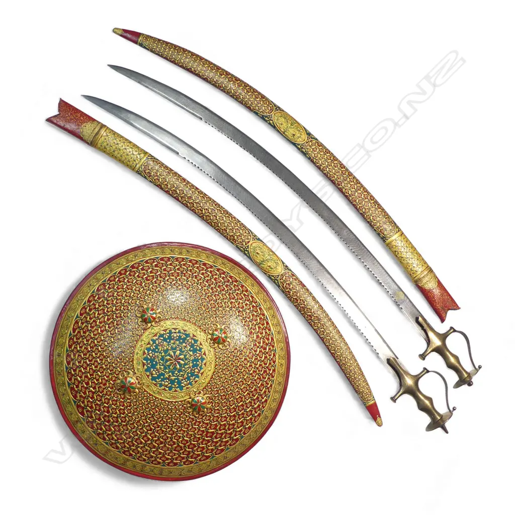 Two Tulwar Swords and matching Dahl Shield from the Punjab Region, India. A matching pair of 28" deeply-curved wootz blades with a gold cartouche to each; double-edged tips and serrated notch-backed to 3/4 of their lengths. The hilts are profusely gold damascened (one with a pommel spike, the other a dome). Matched leather scabbards with raised gilding on a red ground. Along with a matching Dahl shield, dia.20". Believed to have been made as a presentation set. Image 1++